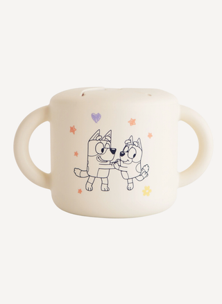A cream-colored Mushie x Bluey silicone snack cup with two handles, featuring Bluey and Bingo dancing with colorful stars and a soft petal-style spill-proof lid.