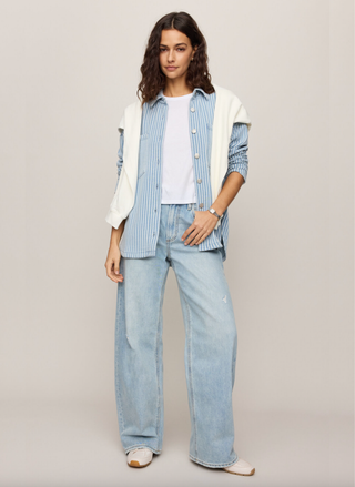Woman wearing a light blue striped shirt, white sweater, and light blue jeans on a plain background