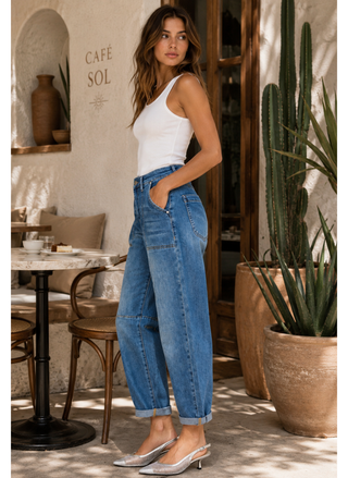 Woman in a white tank top and Risen jeans standing in a casual outdoor setting with plants and a cafe in the background.