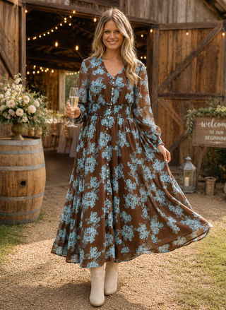Front view of a smiling model wearing the brown and blue floral Talisman Halcyon Dress holding a champagne glass in front of a rustic barn.