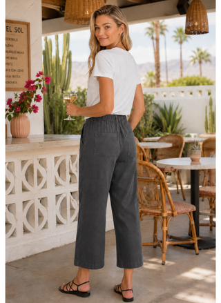 Woman in white top and gray pants standing in a casual outdoor setting with tables and plants.