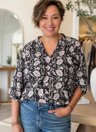 Front view of a smiling woman wearing the black and pink floral Dylan Jolie Cotton Esme Blouse with denim jeans in a boutique setting.