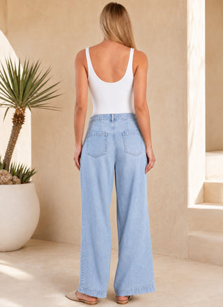 A woman shown from behind wearing light-wash wide-leg denim jeans and a white tank top, standing in a sunlit beige architectural setting with a potted plant nearby.