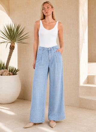 A woman modeling light-wash wide-leg denim jeans with a white tank top, standing in a sunlit architectural setting with a potted plant.