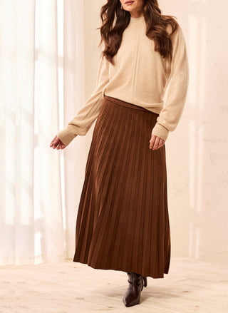 A woman wearing a beige knit sweater and a brown pleated midi skirt, standing in a softly lit room with sheer curtains and light wood flooring.