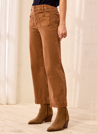 A person wearing brown wide-leg cropped jeans with rolled cuffs and brown ankle boots, standing in a softly lit indoor setting with a light wall and wood floor.