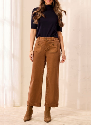 Woman wearing caramel brown TRIBAL Sophia Curvy Straight Ankle Denim paired with a dark top and suede ankle boots, standing in a brightly lit room with white curtains.