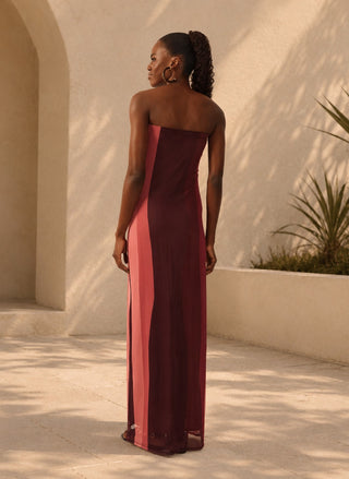 A woman stands outdoors in a warm architectural setting wearing a strapless floor-length gown with vertical color-blocked panels in deep burgundy and dusty rose pink. She is shown from the back, with her hair in a high ponytail and large hoop earrings.