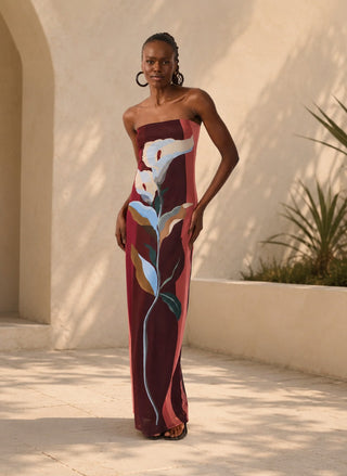 Woman wearing the Dress To Brisa Strapless Tulle Maxi Dress featuring a large calla lily graphic print in burgundy, coral, and soft blue tones. The dress has a fitted silhouette, styled with black strappy heels in an outdoor patio setting with warm sunlight and shadows.
