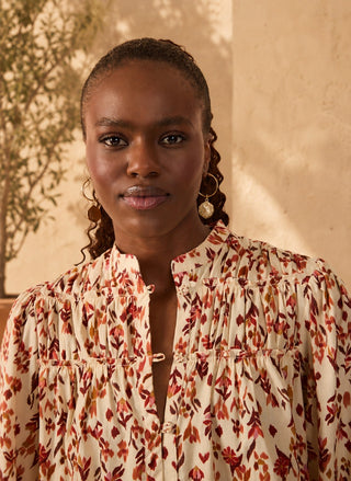 A woman wearing a cream-colored blouse with a rust-red and brown floral pattern, styled with gold hoop earrings, set against a warm beige wall with soft shadows.