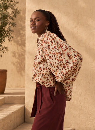 A woman wearing a cream blouse with a rust and brown floral print and dark burgundy wrap skirt, standing in front of a textured beige stucco wall.