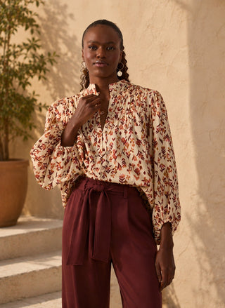 A woman stands outdoors against a warm beige stucco wall wearing a cream blouse with an allover rust-red and brown floral print, paired with high-waisted burgundy trousers with a self-tie waist.