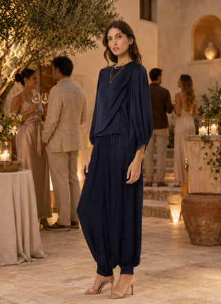 A woman stands in a warmly lit outdoor event space wearing a navy blue draped jumpsuit with voluminous balloon sleeves and ankle-length harem legs, paired with nude high-heeled sandals.