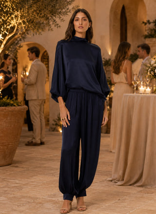 A woman stands in a warmly lit event courtyard wearing a dark navy satin jumpsuit with a high mock neck and voluminous draped sleeves and legs. Guests and tables with candles are softly blurred in the background.