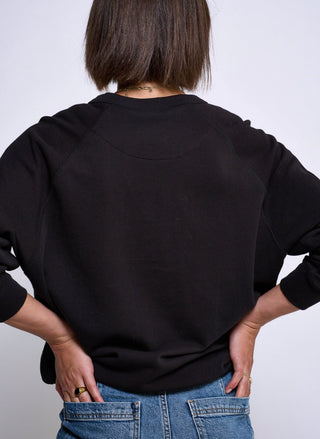 Rear view of a person wearing a black long-sleeve crewneck sweatshirt and blue jeans against a light gray studio background.