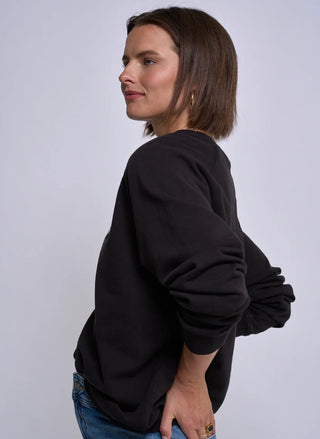 A woman with short brown hair is shown from a side-back angle wearing a black crewneck sweatshirt and blue jeans against a plain light gray studio background.