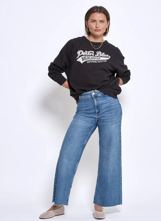 A woman modeling a black crewneck sweatshirt with a white graphic print that reads 'Pura Vida Mexican Style' and blue wide-leg jeans, standing against a plain white studio background.