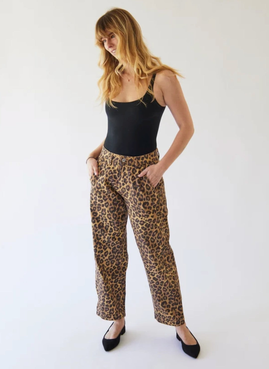 A woman modeling the Porter Blue Tara leopard-print barrel pants with a black camisole top and black flats, standing against a plain white studio background.