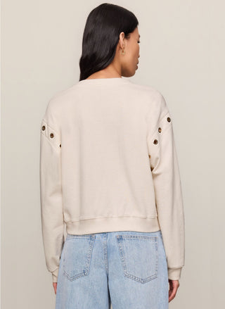 A woman shown from the back wearing a cream-colored cropped sweatshirt with decorative metallic buttons on the shoulders and light blue jeans, set against a plain light beige studio background.