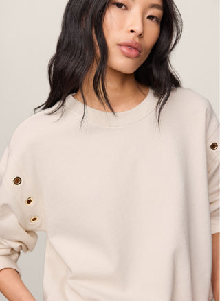 A close-up of a model wearing a cream-colored crewneck sweatshirt with decorative gold eyelet buttons on the sleeves, set against a plain light beige background.