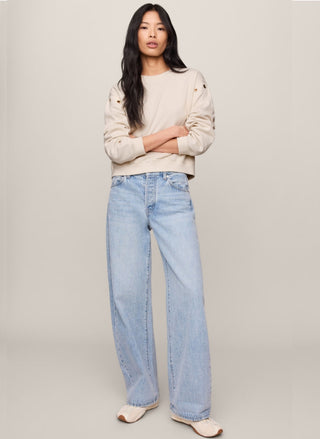 A woman modeling a light beige cropped sweatshirt and wide-leg light blue jeans against a plain light gray studio background.