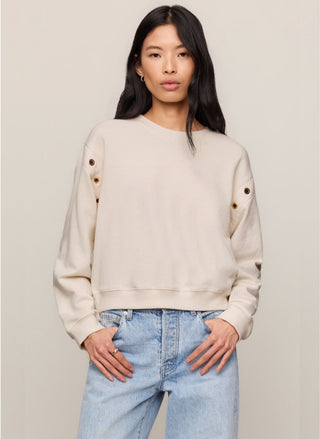 Woman wearing a cream cotton French terry crewneck sweatshirt with gold grommet hardware detail along the shoulders and sleeves, paired with light wash denim.