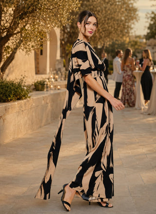 Woman wearing a black and camel abstract print sleeveless jumpsuit with a draped scarf-neck detail and wide-leg silhouette.