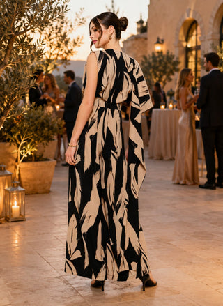 Back view of a woman wearing a black and camel abstract print sleeveless jumpsuit with a draped scarf-neck detail and wide-leg silhouette.