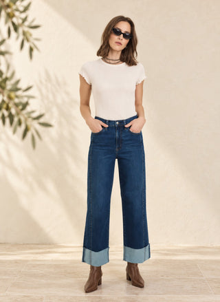 A woman modeling a white short-sleeve top and high-waisted dark blue wide-leg jeans with cuffed hems, paired with brown ankle boots, standing against a textured beige wall with leaf shadows.