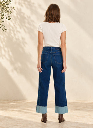 A woman viewed from behind wearing dark blue wide-leg jeans with rolled cuffs, a white short-sleeve top, and brown ankle boots, standing against a warm beige wall with soft leaf shadows.
