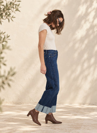 Woman wearing Daze Denim Pleaser high-rise wide-leg cropped jeans in Slow Motion wash, a white tee, brown boot & Sunglasses.