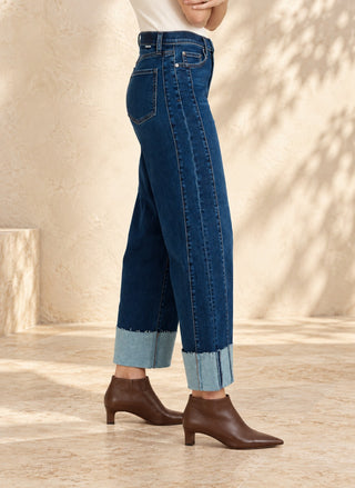 A person shown from the waist down wearing dark blue cropped jeans with rolled light-wash cuffs and brown ankle boots, standing in a sunlit beige interior setting.