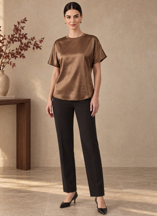 A woman stands in a styled indoor setting wearing a short-sleeve satin-like top in a warm taupe-brown color, paired with black straight-leg pants and black pointed heels.