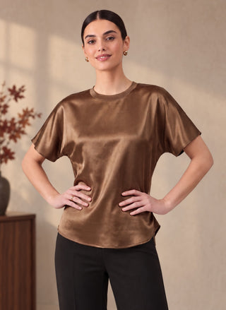 A woman modeling a short-sleeve crewneck top made of shiny brown satin, from August Boutique, paired with black pants, standing against a warm beige wall.