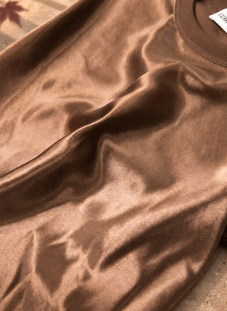 A close-up of a brown satin or silk-like fabric with soft folds and a small white brand tag visible at the collar.