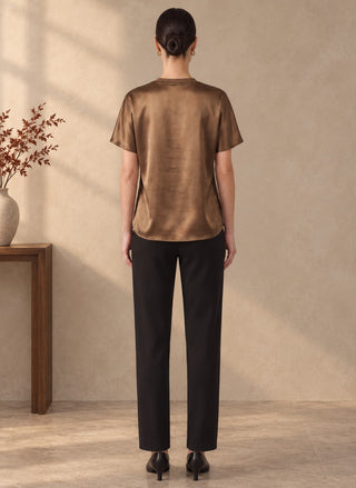 A woman viewed from the back wearing a short-sleeve metallic bronze top and black trousers, standing in a softly lit indoor room with a textured beige wall and a wooden side table with dried branches on the left.
