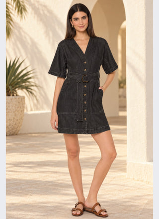 A woman modeling a short-sleeve dark gray denim dress with a V-neck, button front, and matching belt, standing in a sunlit outdoor corridor with arches and potted plants.