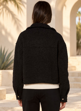 A person viewed from behind wearing a black textured cropped jacket over a cream ribbed top and black pants, standing outdoors in front of a beige building with arches and plants.
