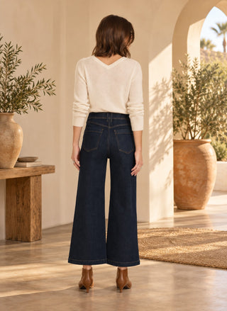 A woman stands with her back to the camera wearing dark blue wide-leg jeans, a cream long-sleeve knit top, and brown ankle boots in a sunlit Mediterranean-style interior with potted plants and arches.
