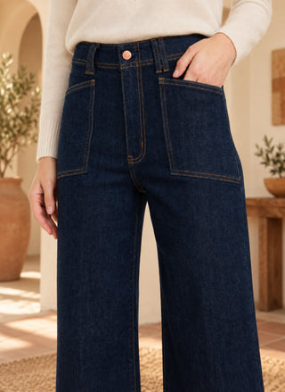 A person wearing dark indigo wide-leg jeans with a cream ribbed sweater, standing in a warm, sunlit indoor setting with plants and wooden furniture.
