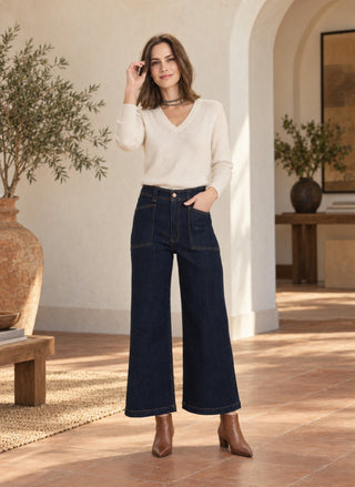 A woman stands in a sunlit architectural interior wearing a cream V-neck long-sleeve top, dark wide-leg denim jeans, and brown ankle boots.