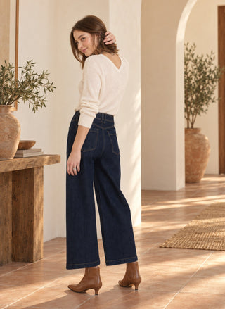 A woman wearing a cream long-sleeve knit top and dark blue wide-leg cropped jeans, paired with brown ankle boots, standing in a warm, sunlit interior with arches and potted plants.
