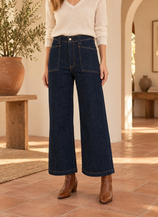 A model wearing DAZE Valley Girl High Rise Patch Pocket Wide Ankle, a cream V-neck sweater, and brown leather ankle boots, standing in a warm, sunlit room with terracotta tile floors and arches.