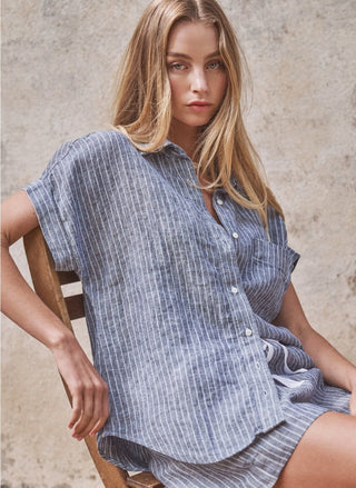 Sideview of the Santa Rosa Linen Short Sleeve Button Shirt in a classic vertical stripe pattern, featuring a button-down placket, short sleeves, and a front chest pocket.  Model is sitting on a chair.