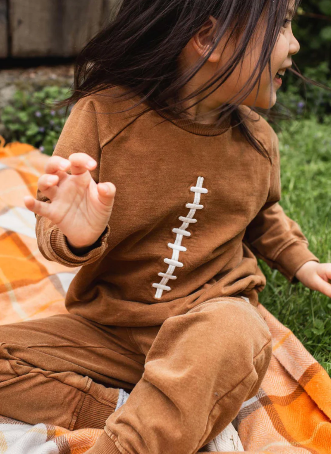 Child wearing a brown onesie with a white design, sitting on grass.