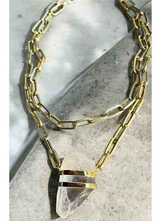 Raw clear quartz pendant set in solid bronze hanging from a double layer gold-toned paperclip chain on a white top.