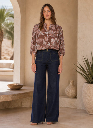 A woman stands in a sunlit architectural setting wearing a brown floral long-sleeve blouse tucked into dark wide-leg denim jeans.