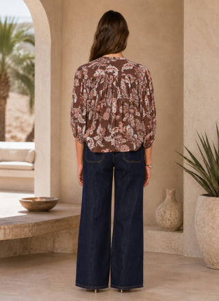 A woman stands with her back to the camera wearing a brown floral long-sleeve blouse and dark wide-leg jeans in a warm beige architectural setting.