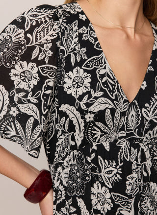 A close-up of a woman wearing a black V-neck dress with a bold white floral and paisley print. The shot includes her neck, chest, and one arm with a dark red bangle.