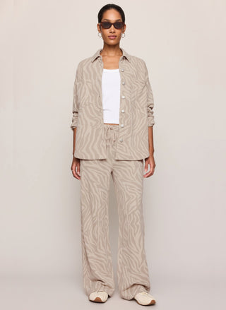 A model wearing a beige and white zebra-print linen lounge set, consisting of a button-up shirt and matching wide-leg pants, styled with a white tank top, sunglasses, and slip-on shoes against a neutral studio background.